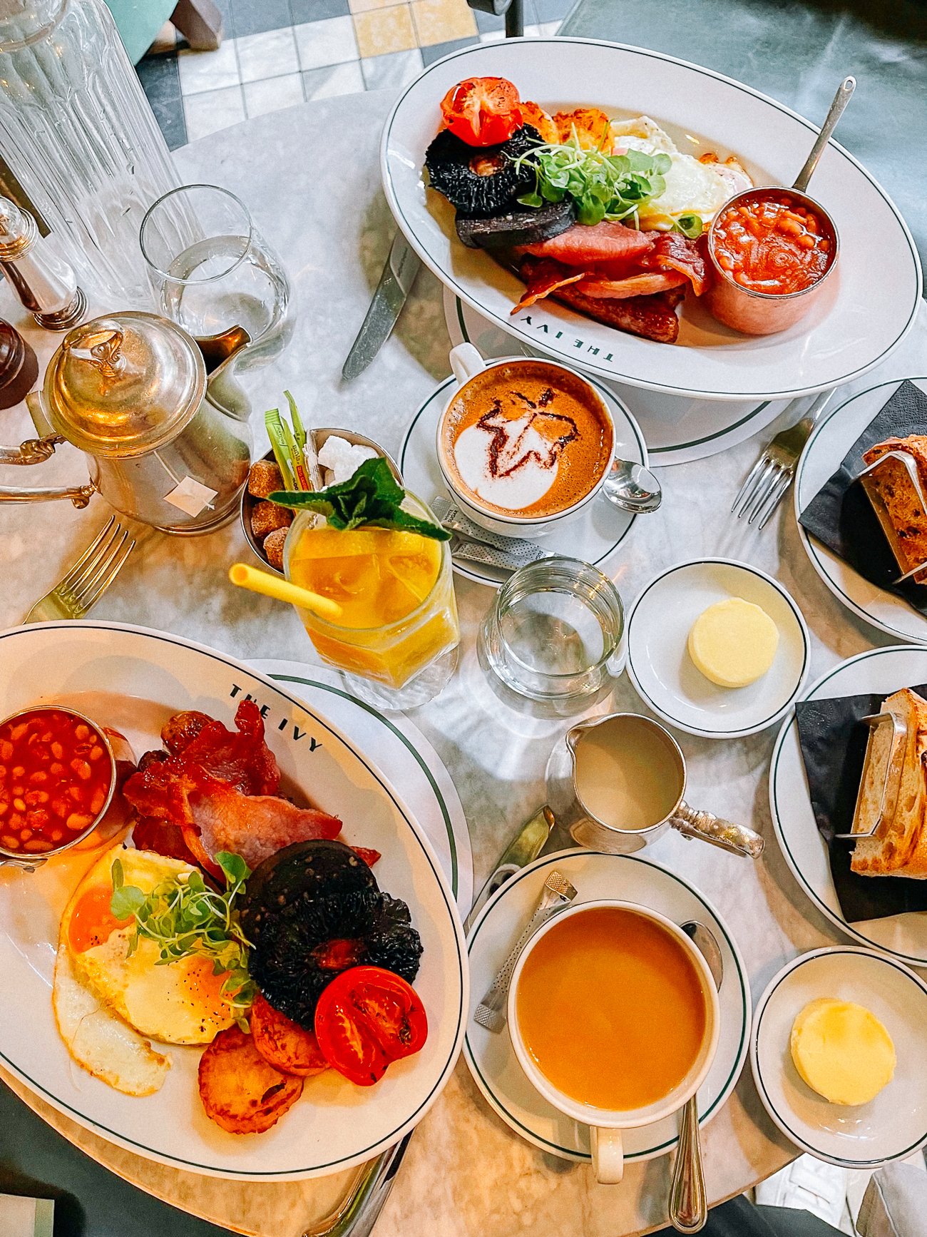 Overhead shot of two English breakfast spreads of beans, bacon, eggs, tomatoes, mushroom, toast and butter with cappuccinos and yellow juice