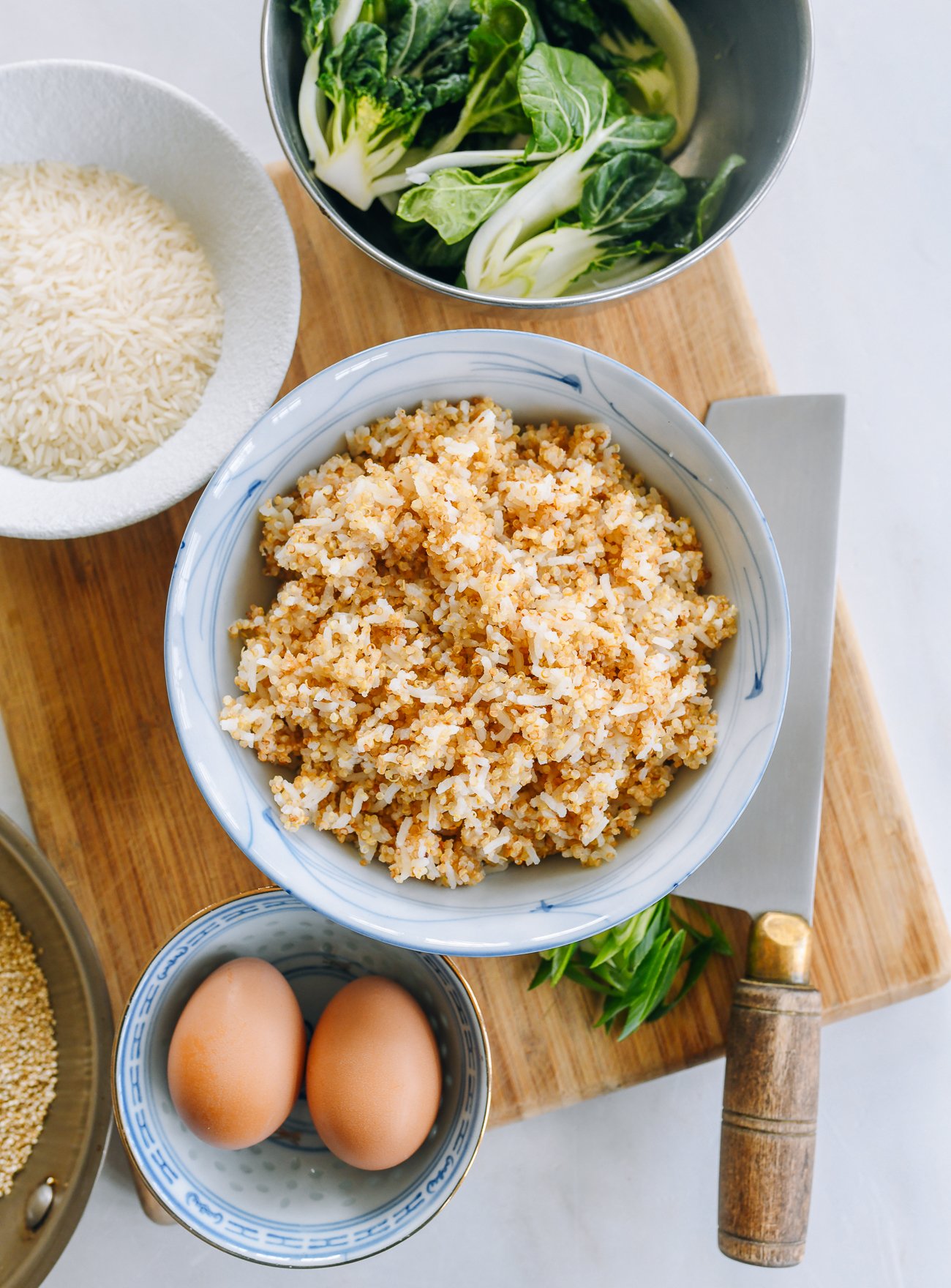 cooked quinoa rice mix on a cutting board with eggs, vegetables and bowls of uncooked rice and quinoa
