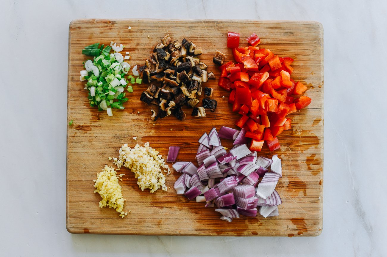 Cutting board with piles of diced red bell pepper, shiitake mushrooms, red onion, scallions, and minced ginger and garlic