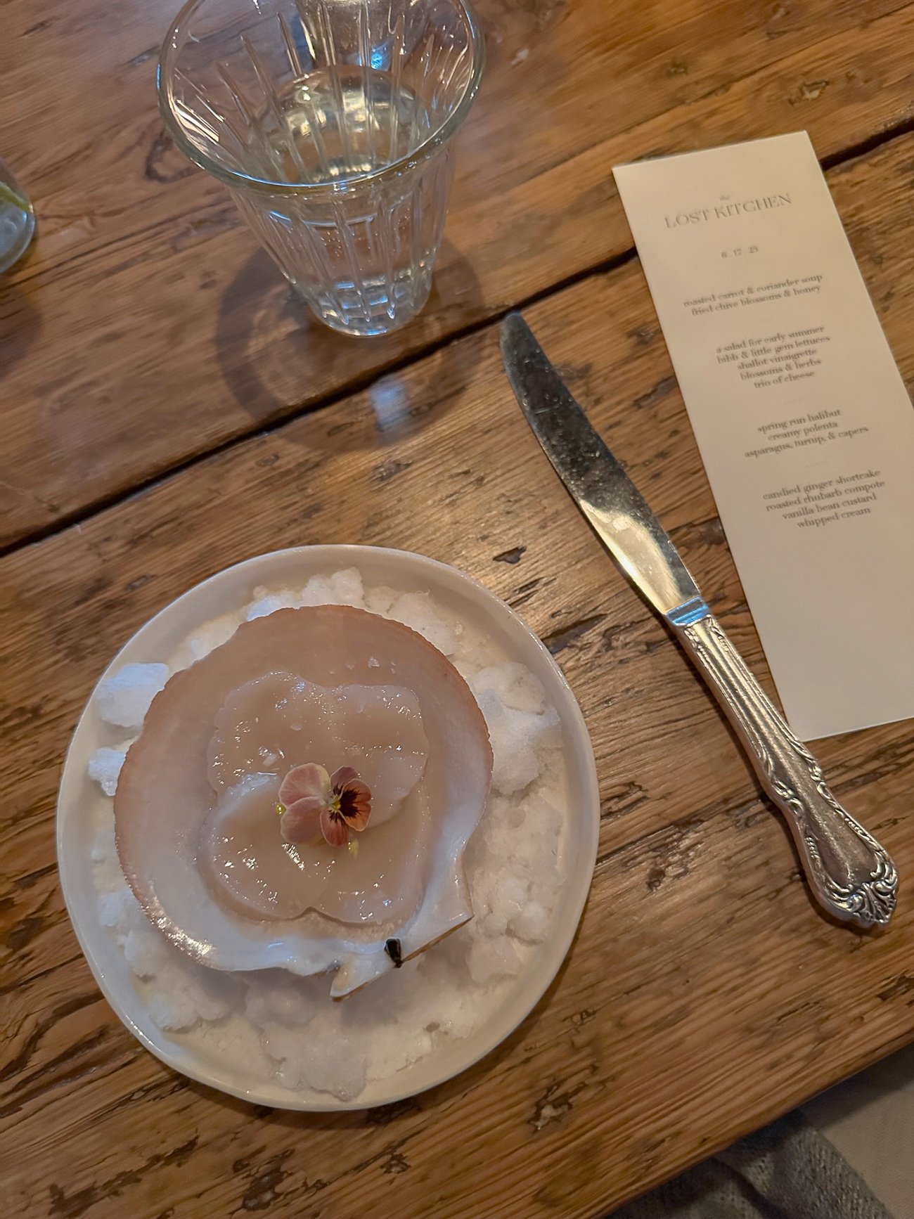 Sliced scallop in shell with a purple flower on a wooden table at The Lost Kitchen
